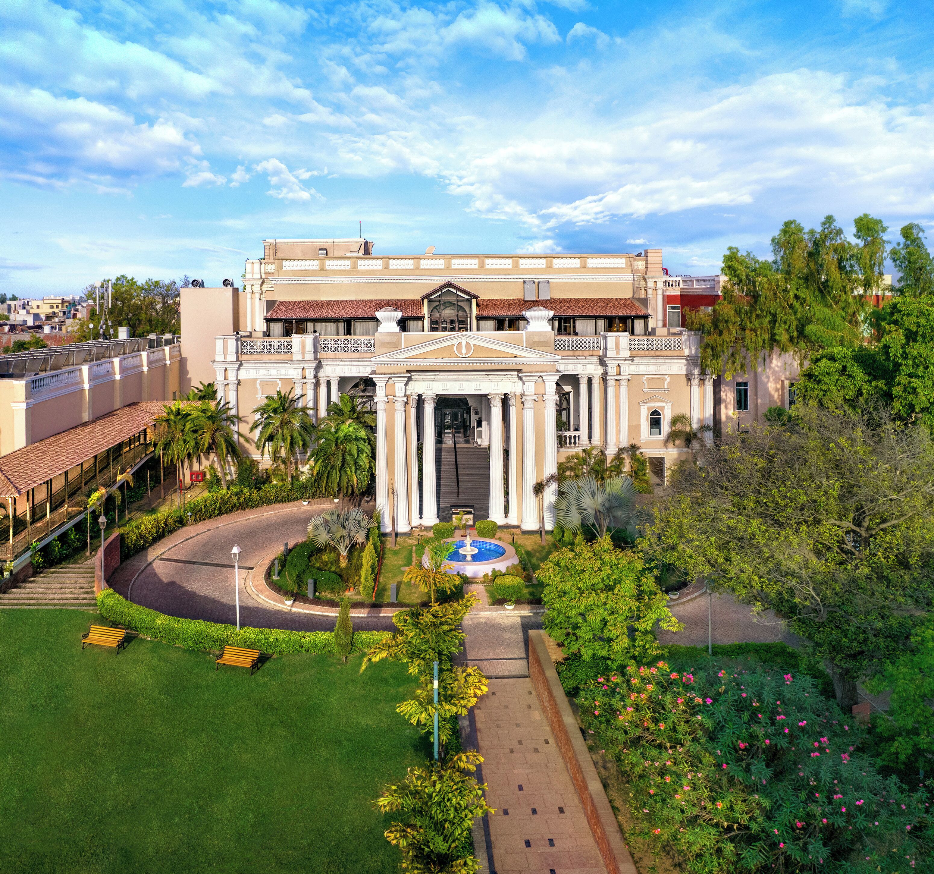 Un edificio grande con una entrada central, rodeado de un jardín cuidado, palmeras y una piscina.