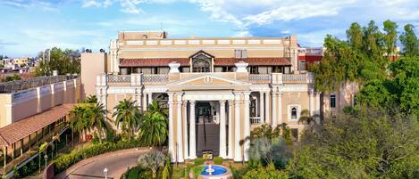 Un edificio grande con una entrada central, rodeado de un jardín cuidado, palmeras y una piscina.
