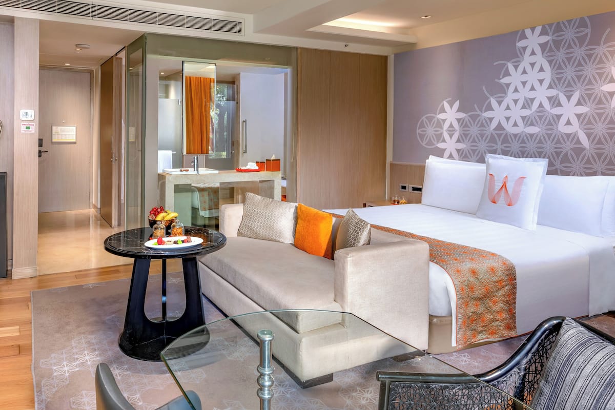 A modern hotel room with a bed, a sofa, a glass coffee table, and a bathroom visible through an open door.
