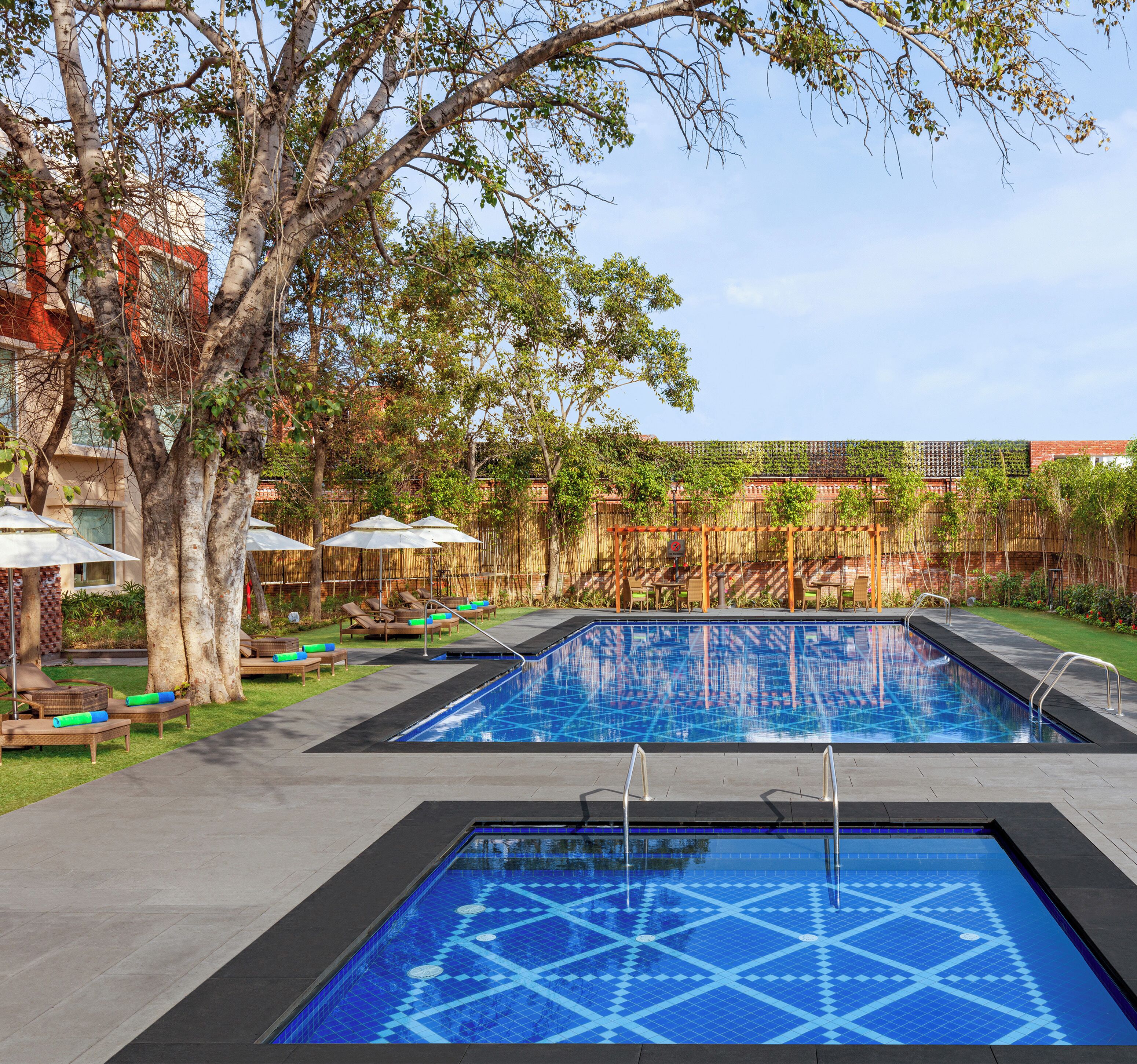 Una piscina con un diseño geométrico de azulejos, rodeada de sillas de descanso y sombrillas, con árboles y un edificio al fondo.