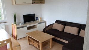 A modern living room with a wooden coffee table, a dark brown sofa with beige cushions, a flat-screen TV on a white cabinet, and a small plant on the side table.