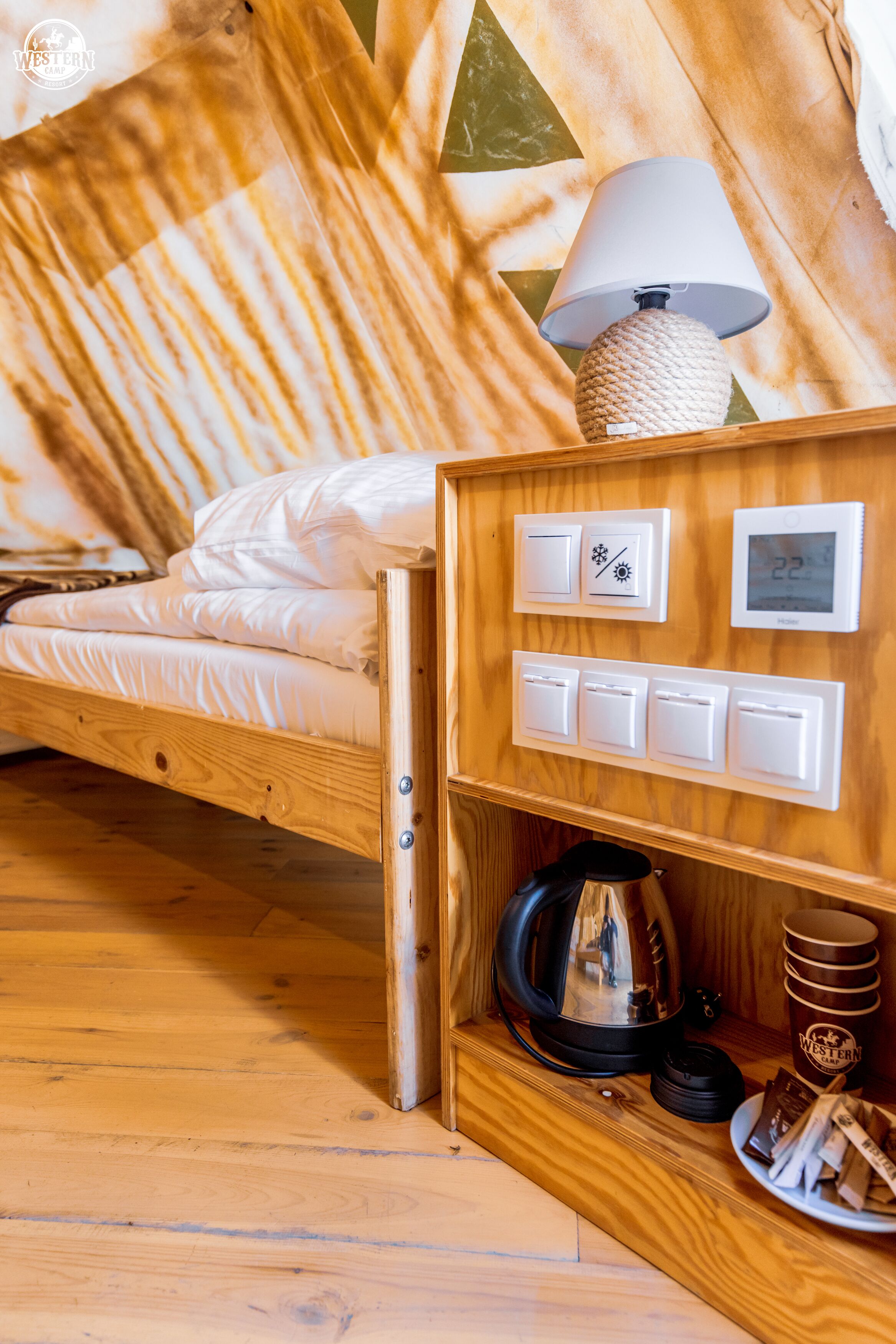 A wooden bed with a white mattress, a lamp, a thermostat, and a shelf with a kettle and a mug.
