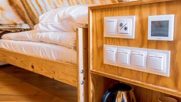 Una cama de madera con colchón blanco, una lámpara, un termostato y un estante con una tetera y una taza.