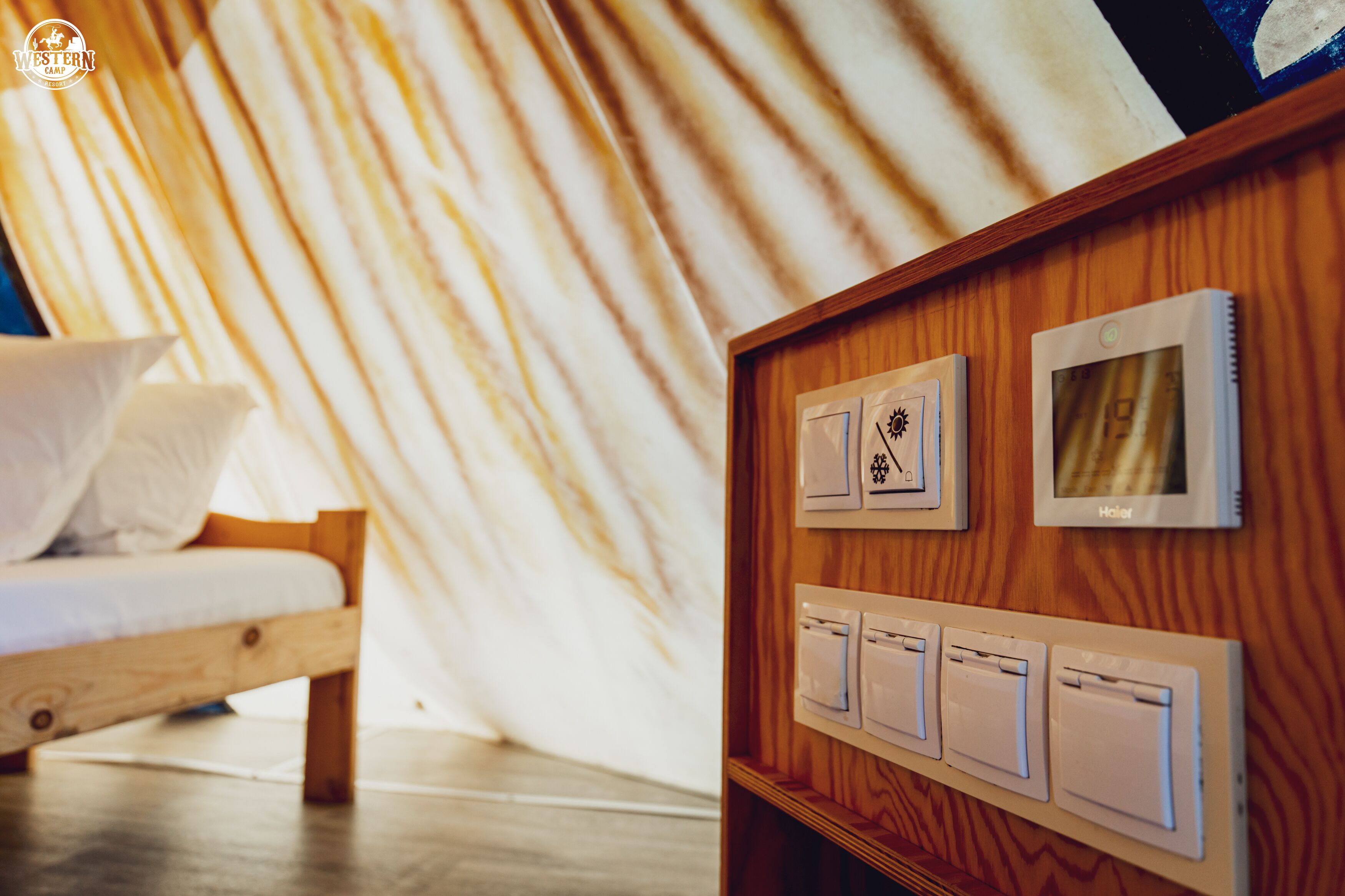 Una cama de madera con colchón y almohadas blancas, una pared de madera con un panel de madera, y un termostato con una pantalla pequeña.