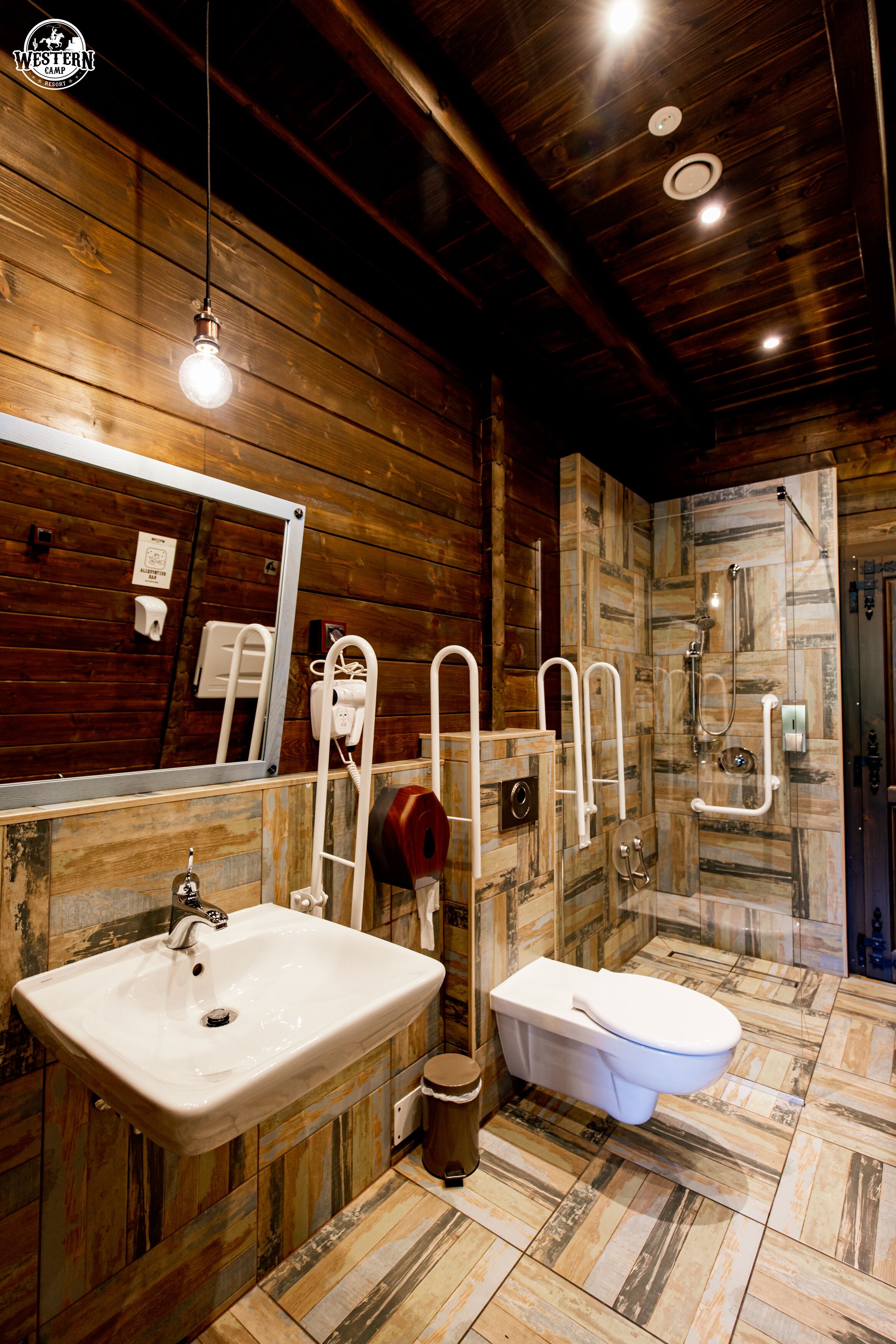 Ein rustikales Badezimmer mit Holzwand, großem Spiegel, Waschbecken, Toilette und Duschbereich.
