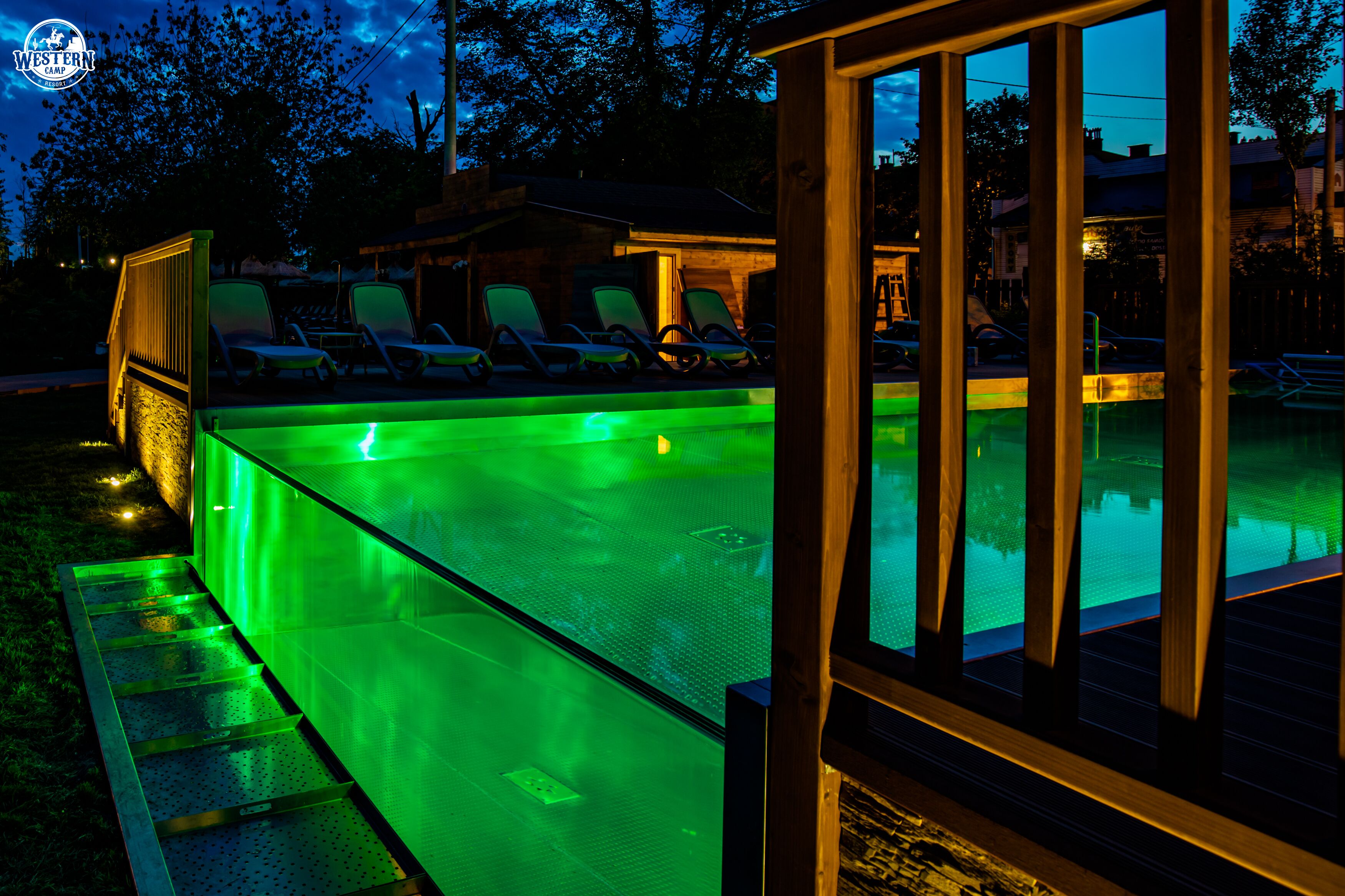 A swimming pool with a green-lit edge, surrounded by lounge chairs and a wooden deck.
