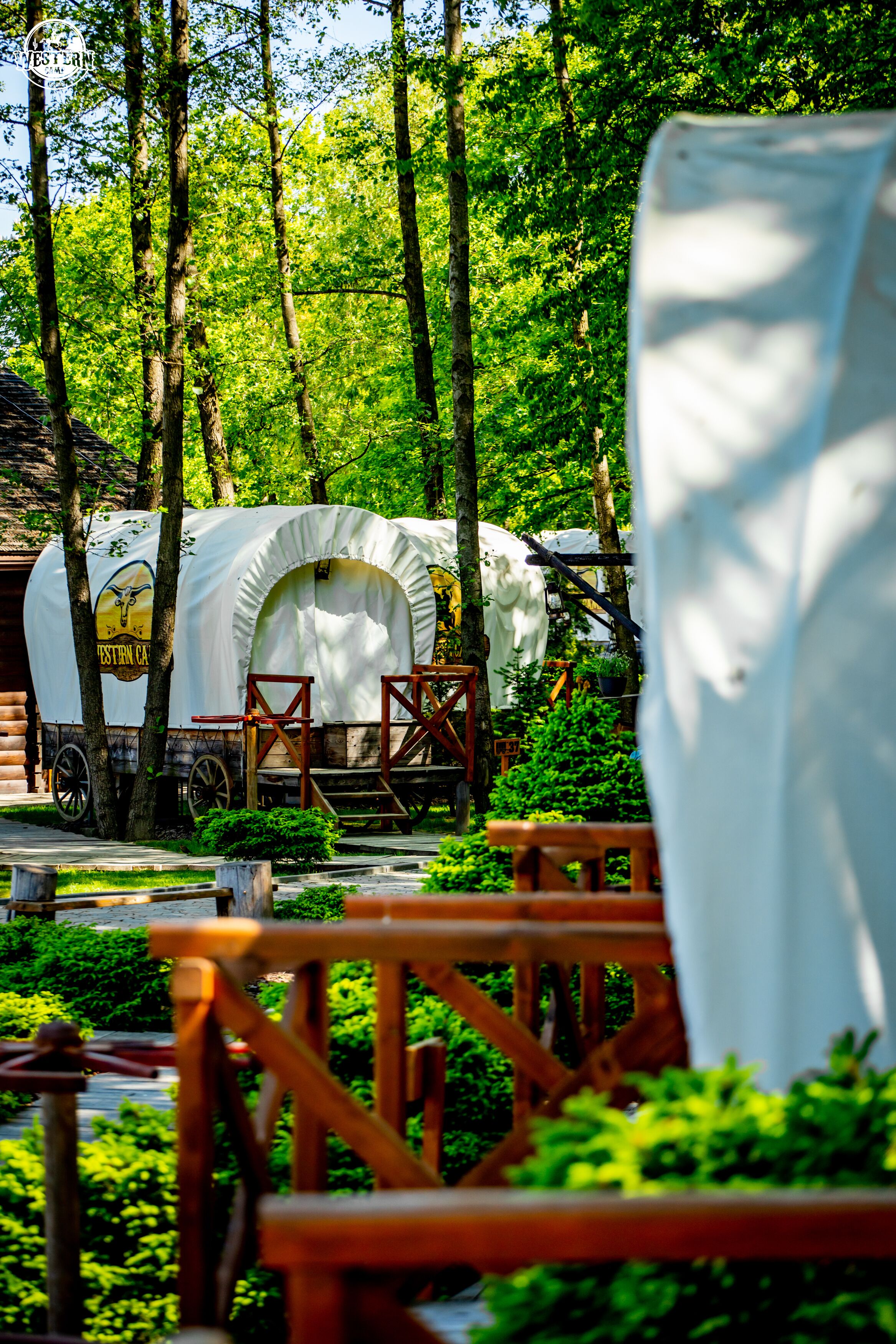 A covered wagon with a logo, surrounded by trees and wooden structures in a park setting.