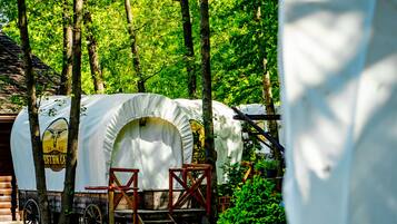 A covered wagon with a logo, surrounded by trees and wooden structures in a park setting.