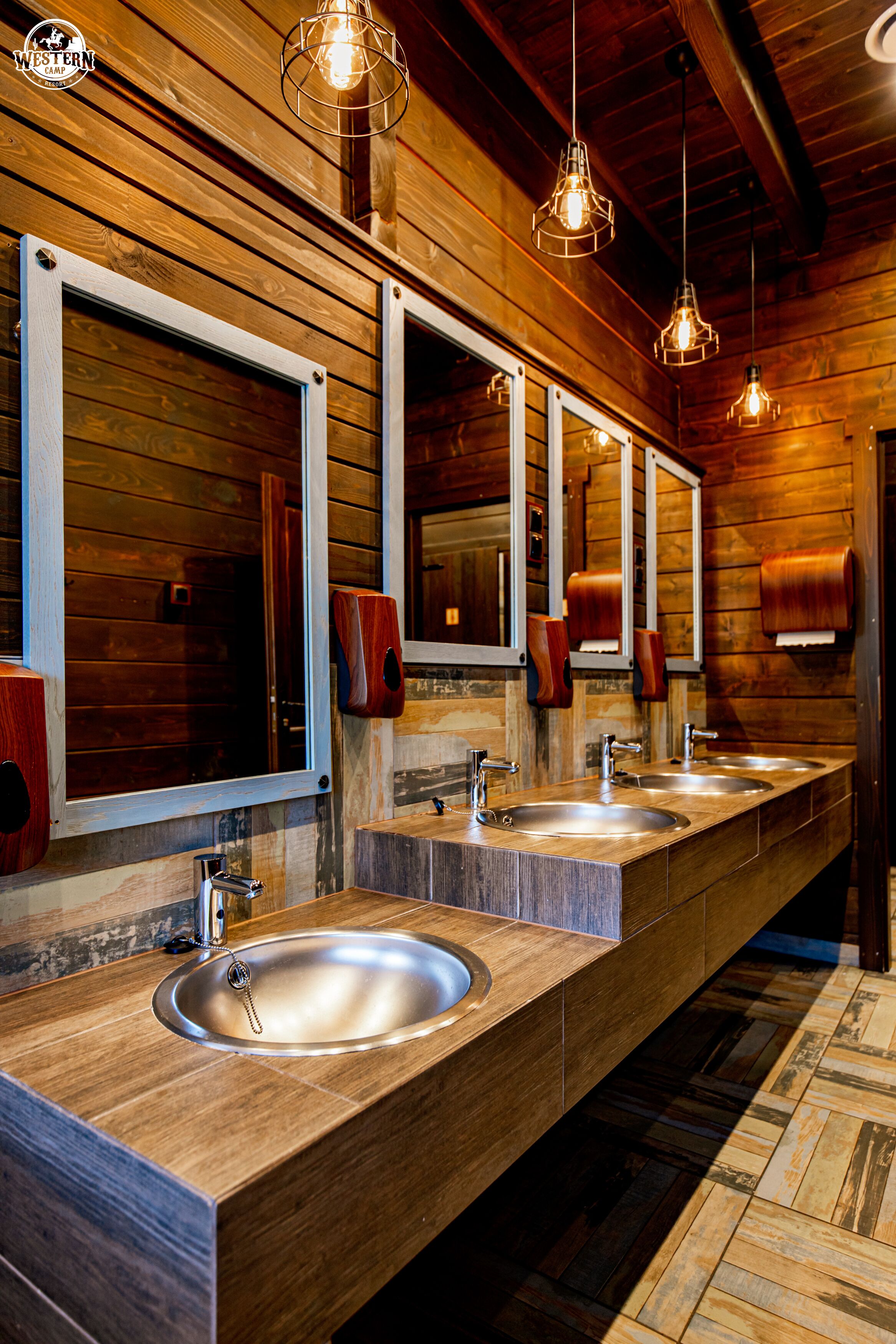 A long, wooden bathroom with three sinks, wooden walls, and pendant lights.