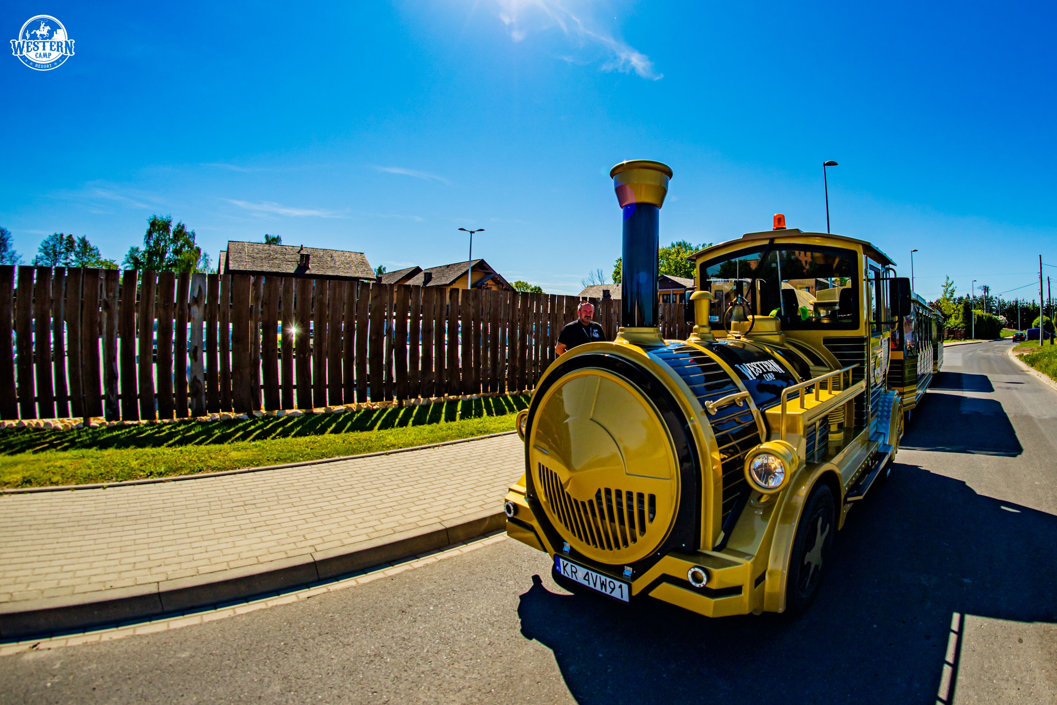 A yellow train on a paved road with a wooden fence and greenery in the background.