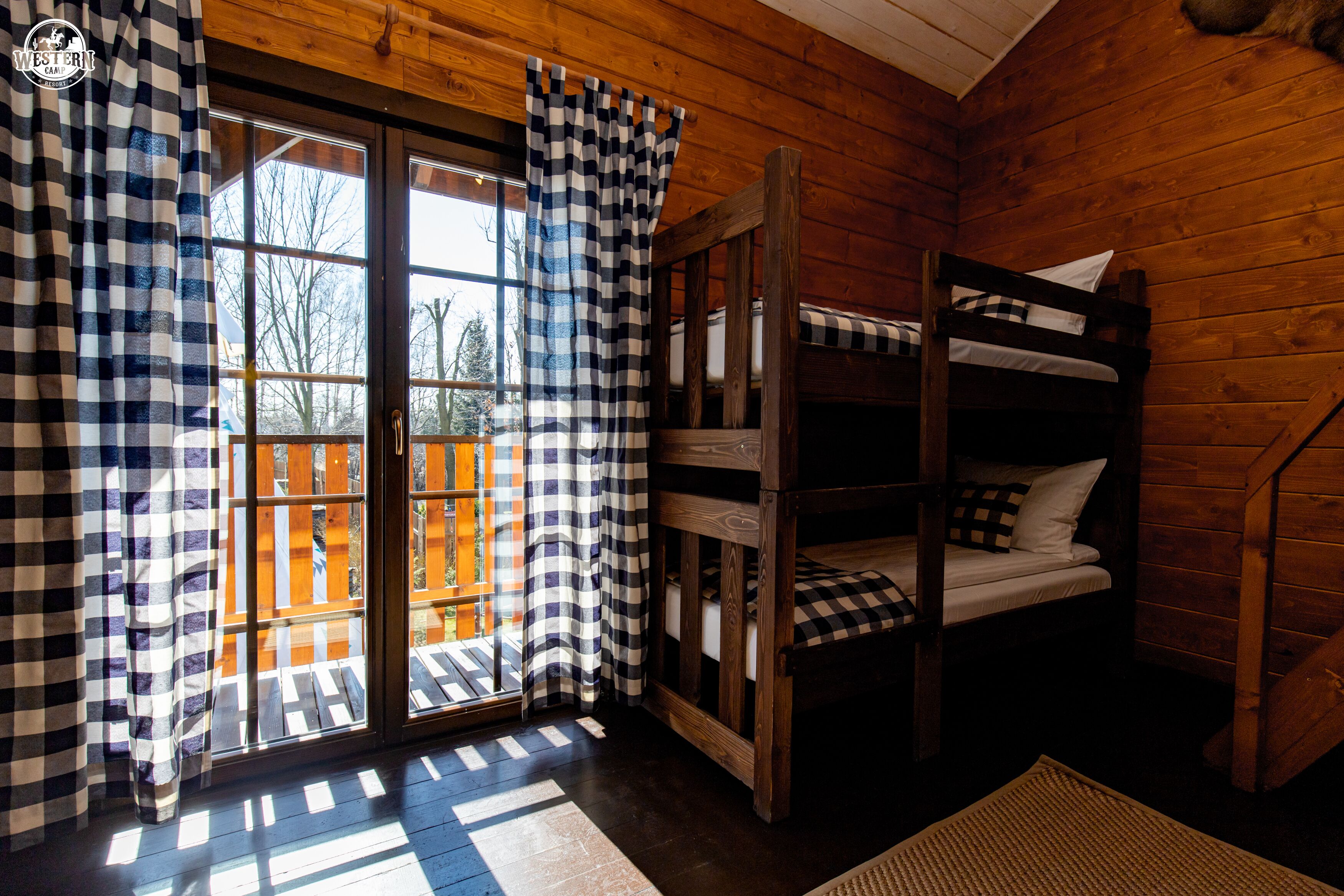 Una habitación tipo cabaña con una cama litera de madera, cortinas a cuadros y vistas a través de un ventanal.