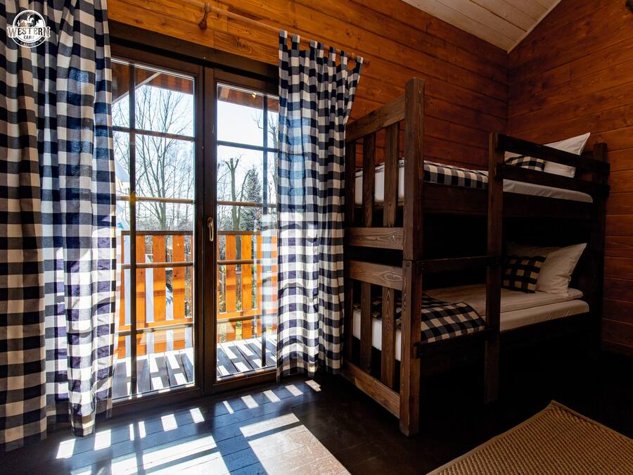 Una habitación tipo cabaña con una cama litera de madera, cortinas a cuadros y vistas a través de un ventanal.