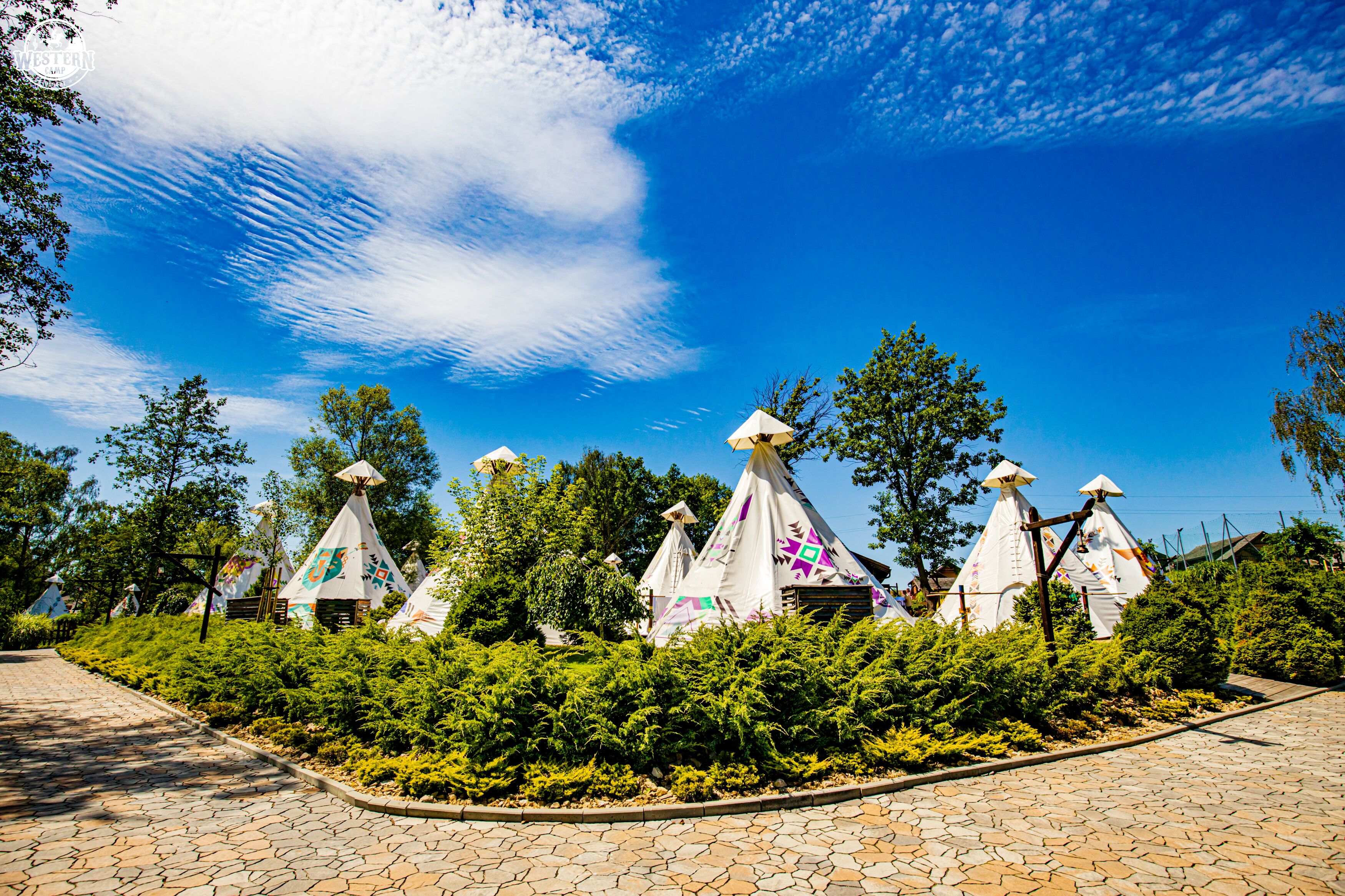 A paved pathway leading to a row of white tents with colorful patterns, surrounded by lush greenery.