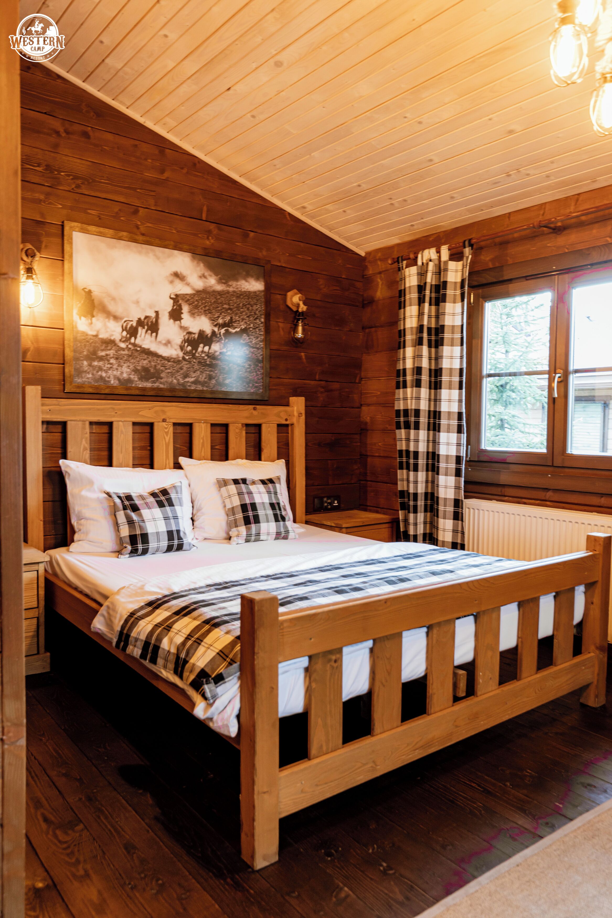 A wooden cabin room with a bed, a mounted picture, and a window with curtains.