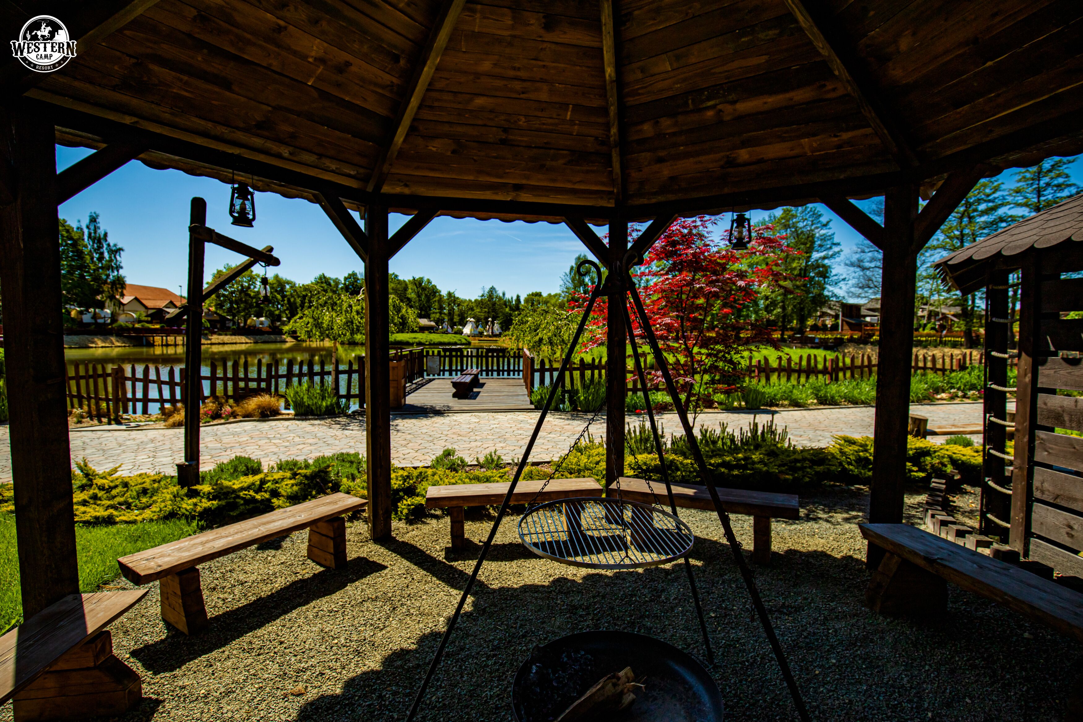 A wooden gazebo with benches and a swing, overlooking a garden with a pond and a wooden fence.