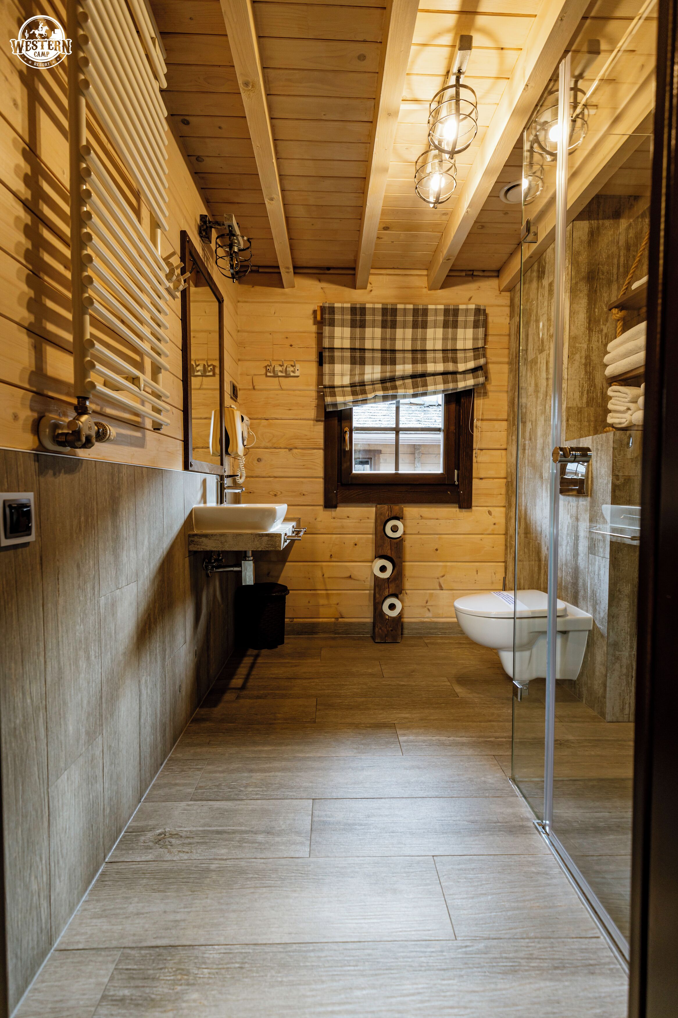 A narrow, wooden-paneled bathroom with a sink, toilet, and a glass-enclosed shower area.