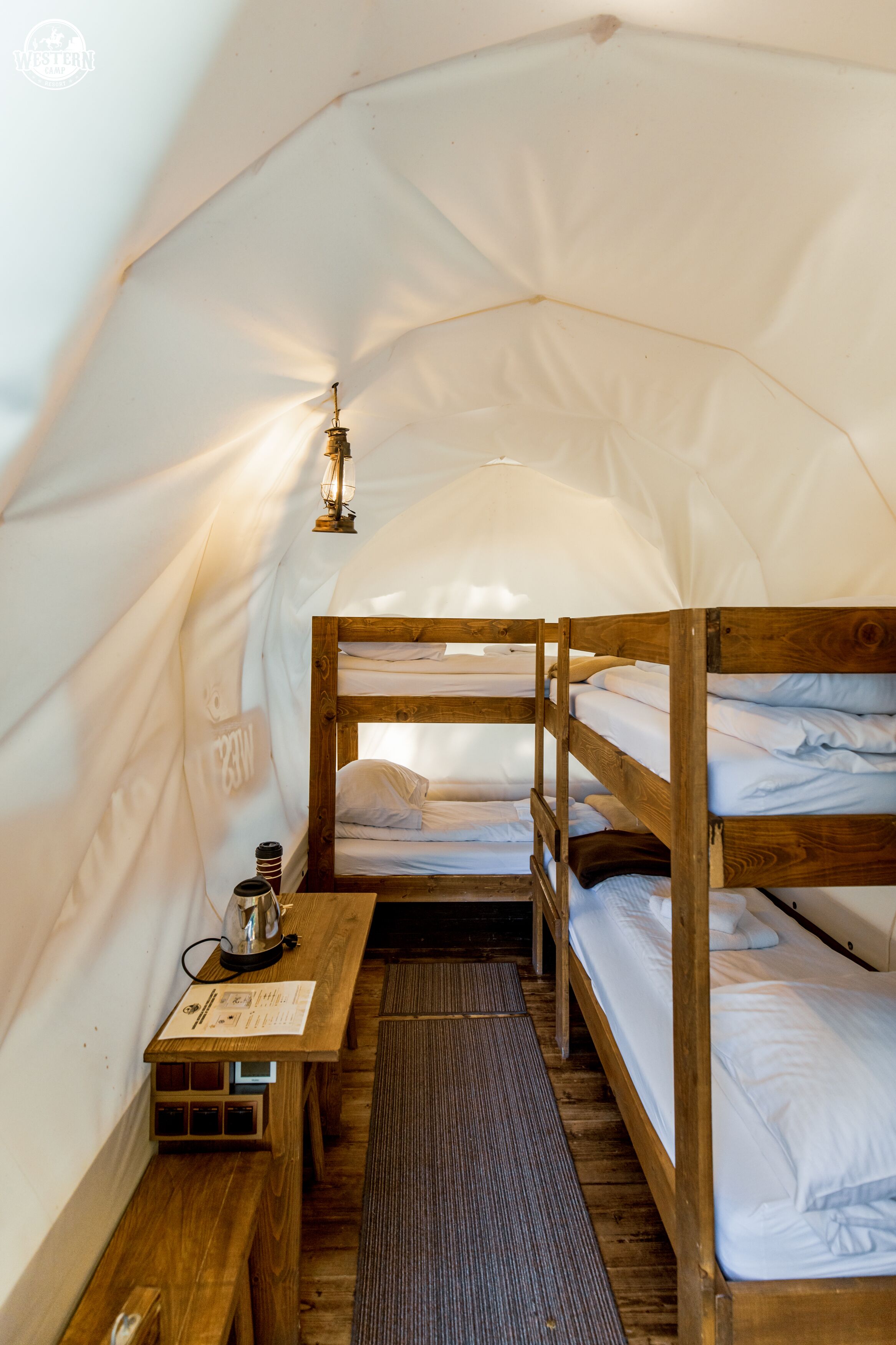 Een tent met stapelbedden, een tafel met waterkoker en een lantaarn.