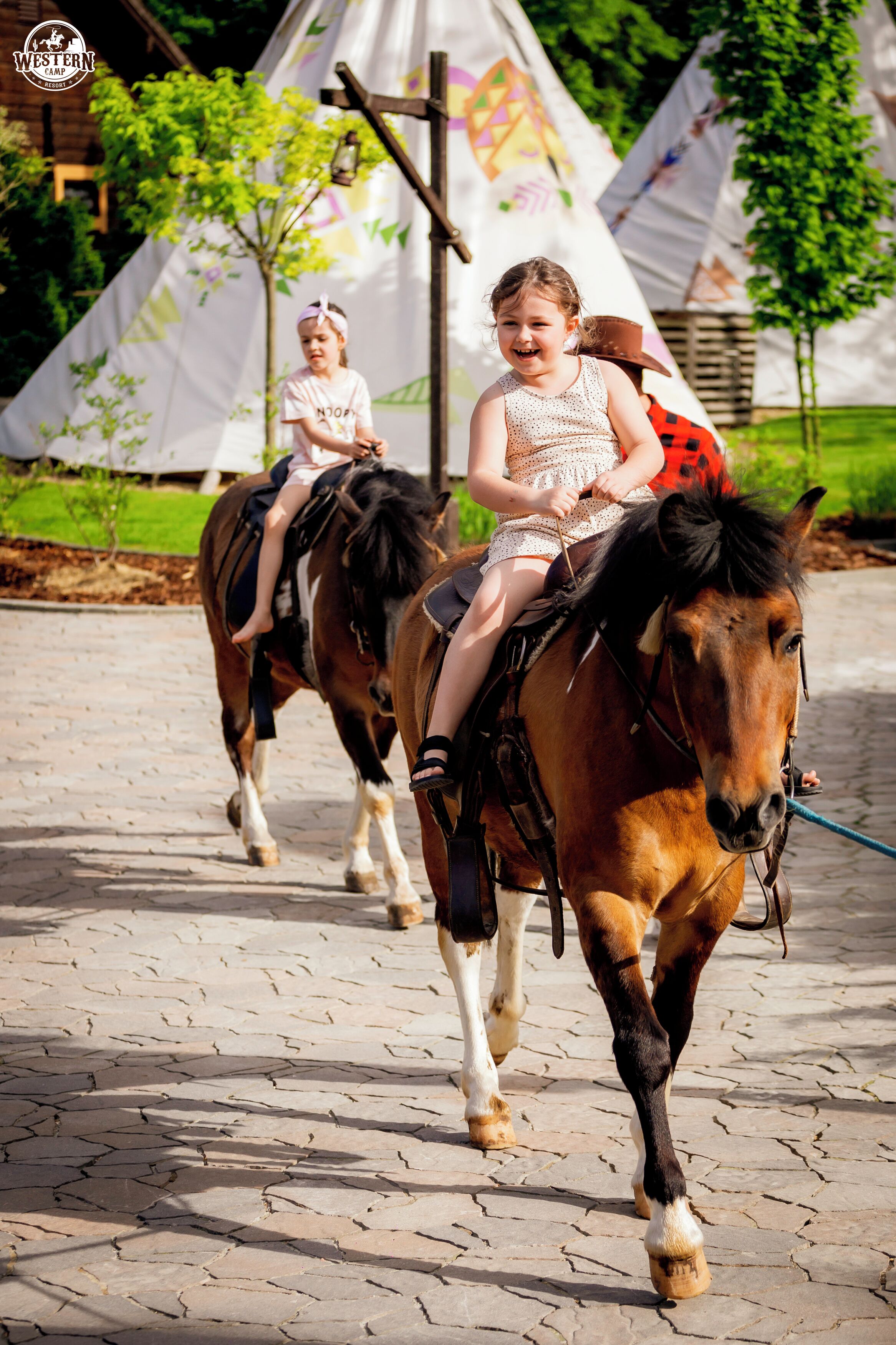 Two children riding horses on a paved path with tents in the background.