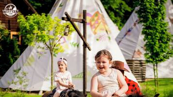 Two children riding horses on a paved path with tents in the background.