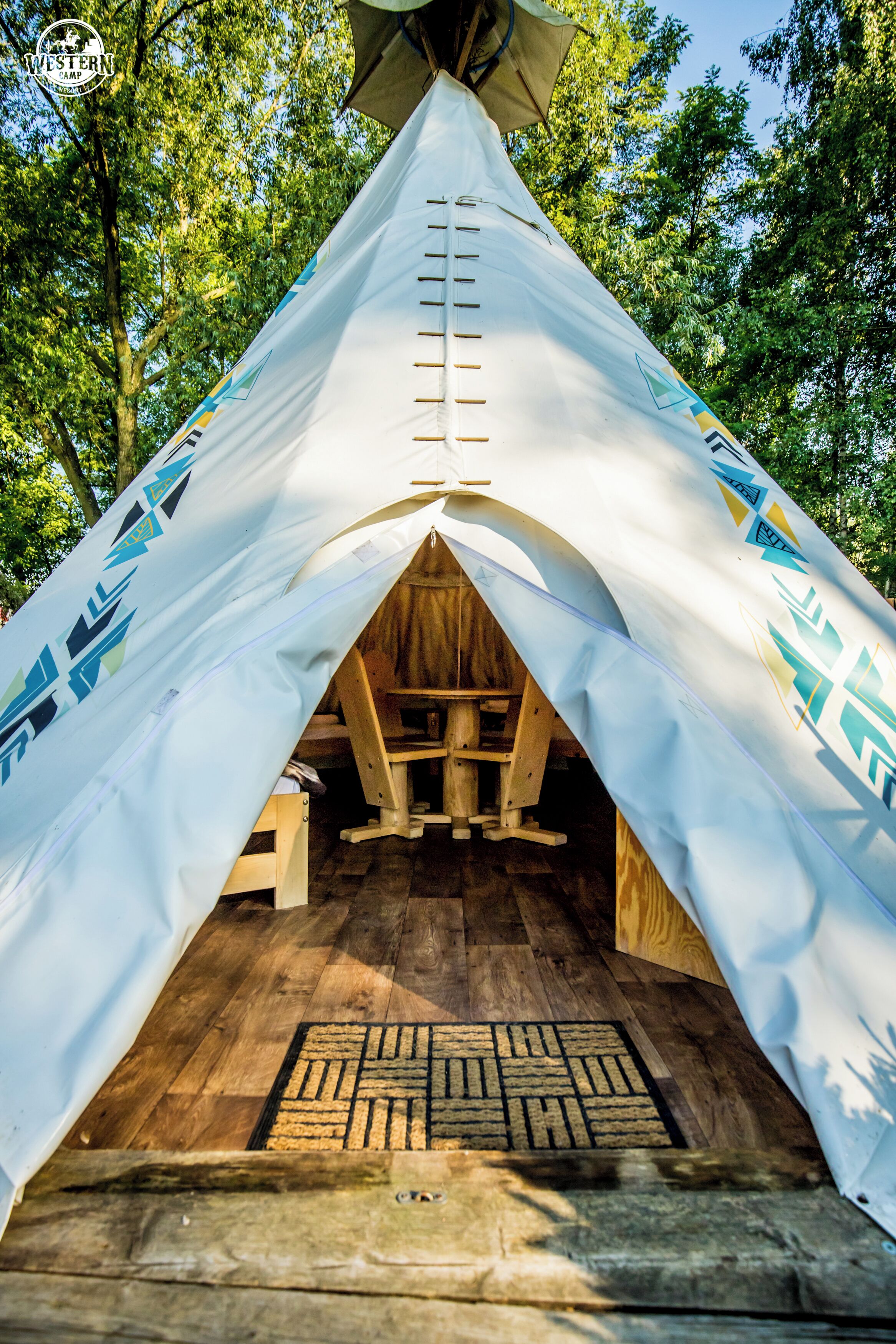 A teepee with a ladder leading to the entrance, wooden flooring inside, and a door mat at the base.