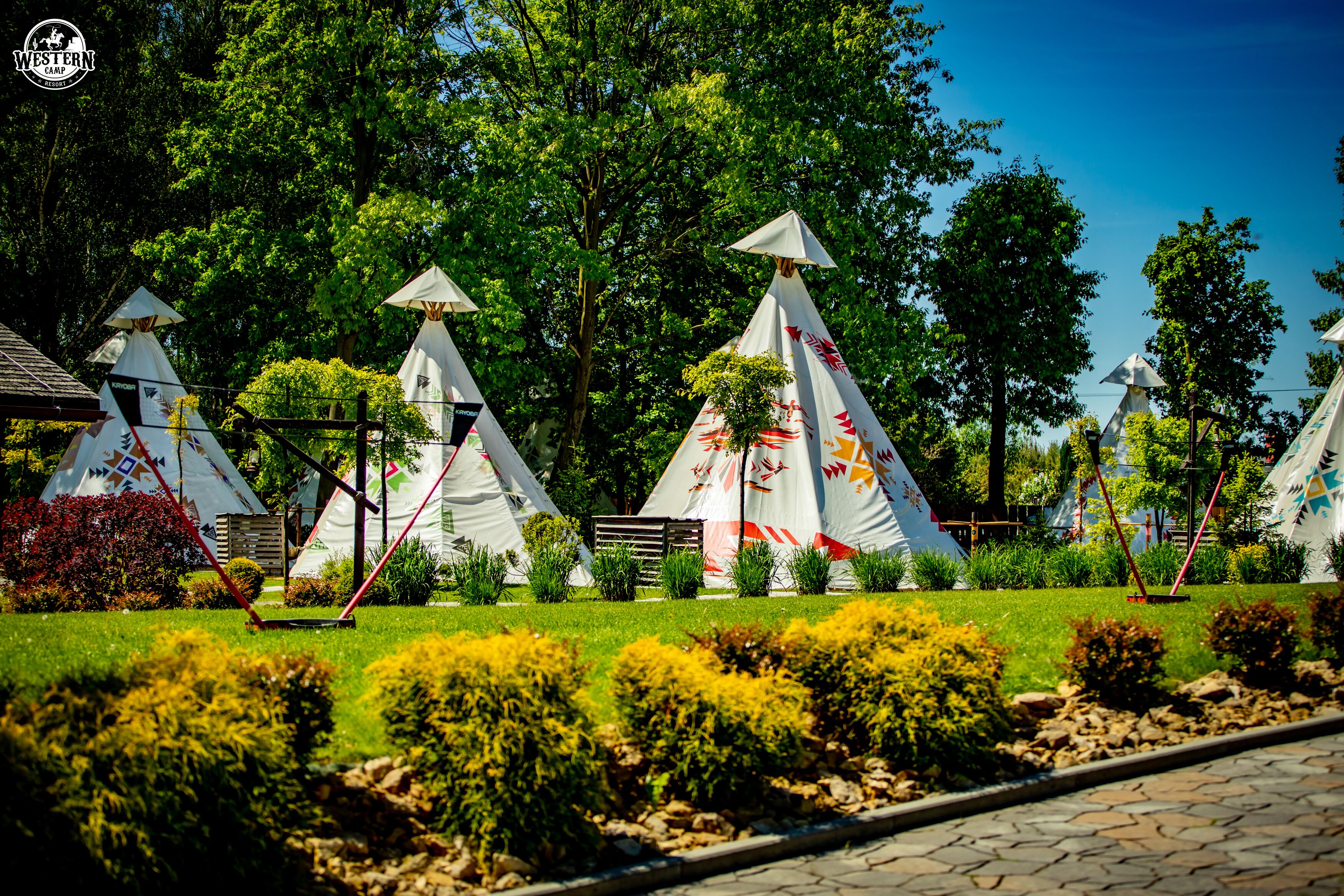 A campsite with multiple white tents with conical roofs, surrounded by lush greenery and trees.