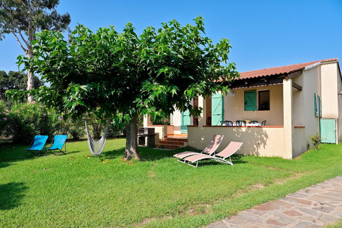 A house with a garden, including a hammock, blue chairs, and a tree.