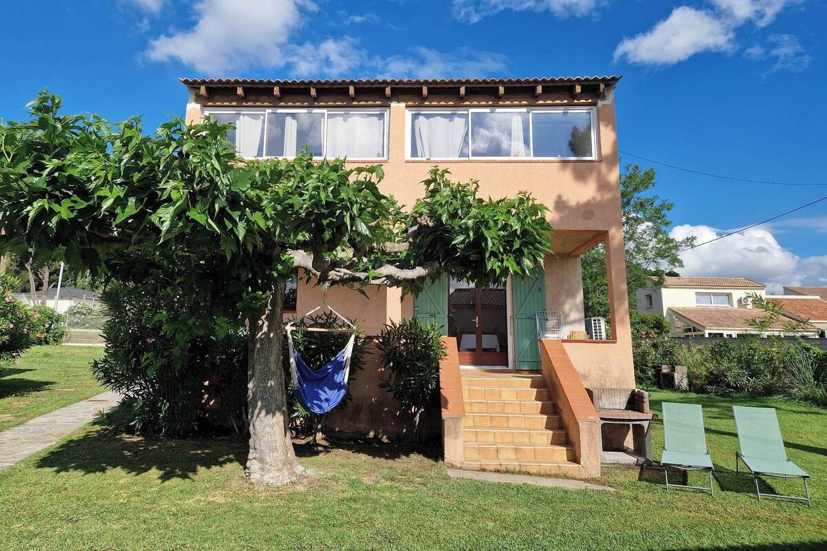 A two-story house with a tiled roof, a front porch with steps, and a hammock hanging from a tree.
