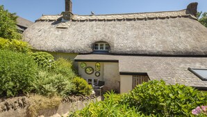 A traditional thatched-roof house with a small window and a door, surrounded by lush greenery. - Higher Collaton Cottage (Kingsbridge)
