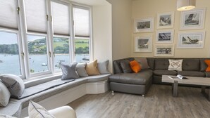 Un salon moderne doté d’une grande fenêtre donnant sur un plan d’eau, d’un canapé d’angle gris, d’un fauteuil blanc et d’une table basse.