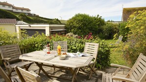 An outdoor patio with a wooden table and chairs, surrounded by lush greenery and a view of houses in the background. - Cindy's Retreat (Kingsbridge)