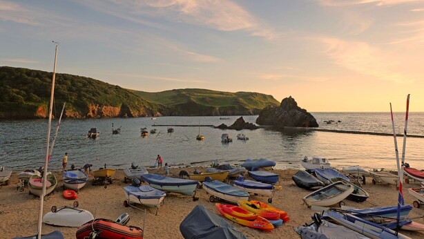 A beach with numerous small boats and canoes, a sandy shore, and a rocky outcrop in the background. - Cindy's Retreat (Kingsbridge)