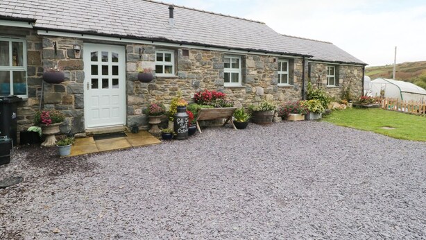 A stone building with a gravel driveway, a white door, and multiple flower pots and planters. - Gelli Glud (Colwyn Bay)
