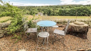 Une table ronde entourée de quatre chaises, située dans un jardin, avec un mur en pierre et une maison visible au loin.