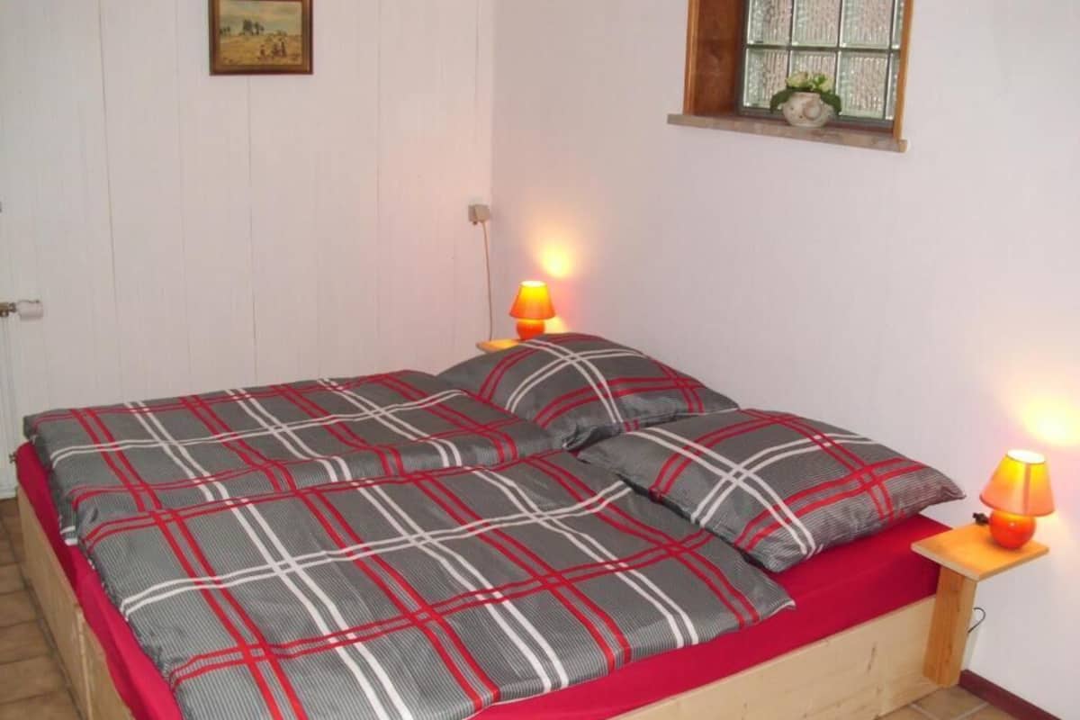 A bed with a plaid bedspread and pillows, two bedside lamps, a window with a view, and a wall-mounted picture.