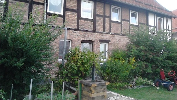 A two-story house with a brick facade, multiple windows, and a garden area with plants and a lawnmower. - Ferienwohnung Langelsheim (Langelsheim)