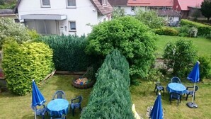 A well-maintained garden with a variety of shrubs and a tall coniferous tree. There are several outdoor tables and chairs, some with blue umbrellas. - Ferienwohnung Langelsheim (Langelsheim)