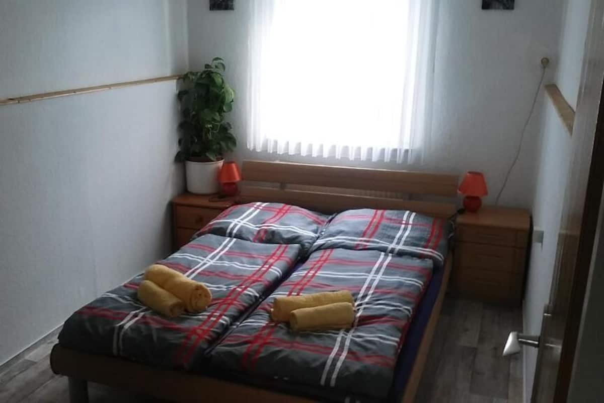 A small, neatly arranged bedroom with a bed, two bedside tables, a potted plant, and a window with curtains.