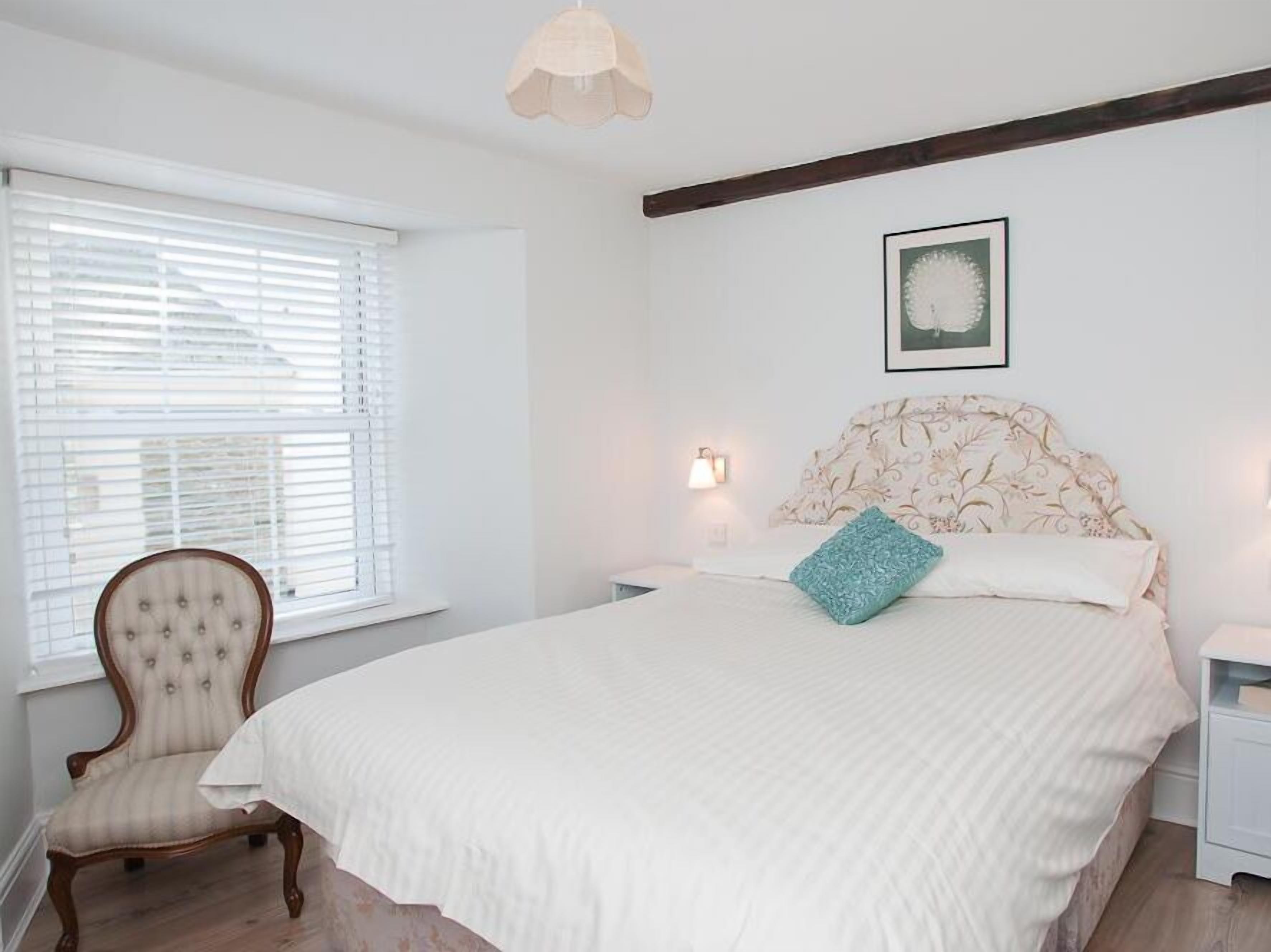 A bedroom with a large bed, a chair, a bedside table, a lamp, and a window with blinds.