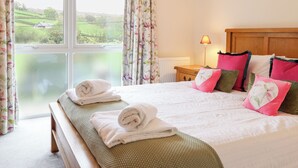 A bedroom with a large window, a bed with rolled towels, a bedside table with a lamp, and a view of a green landscape.