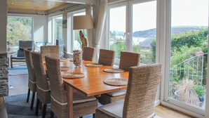A dining area with a wooden table and wicker chairs, large windows with a view of greenery and hills, and a lamp on the table. - Dolphins (Salcombe)