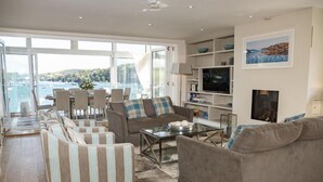 Ein großzügiger Wohnbereich mit einer Couch, Sesseln und einem Couchtisch. Ein Fernseher und ein Kamin sind vorhanden. Das Zimmer hat große Fenster mit Blick auf das Meer.