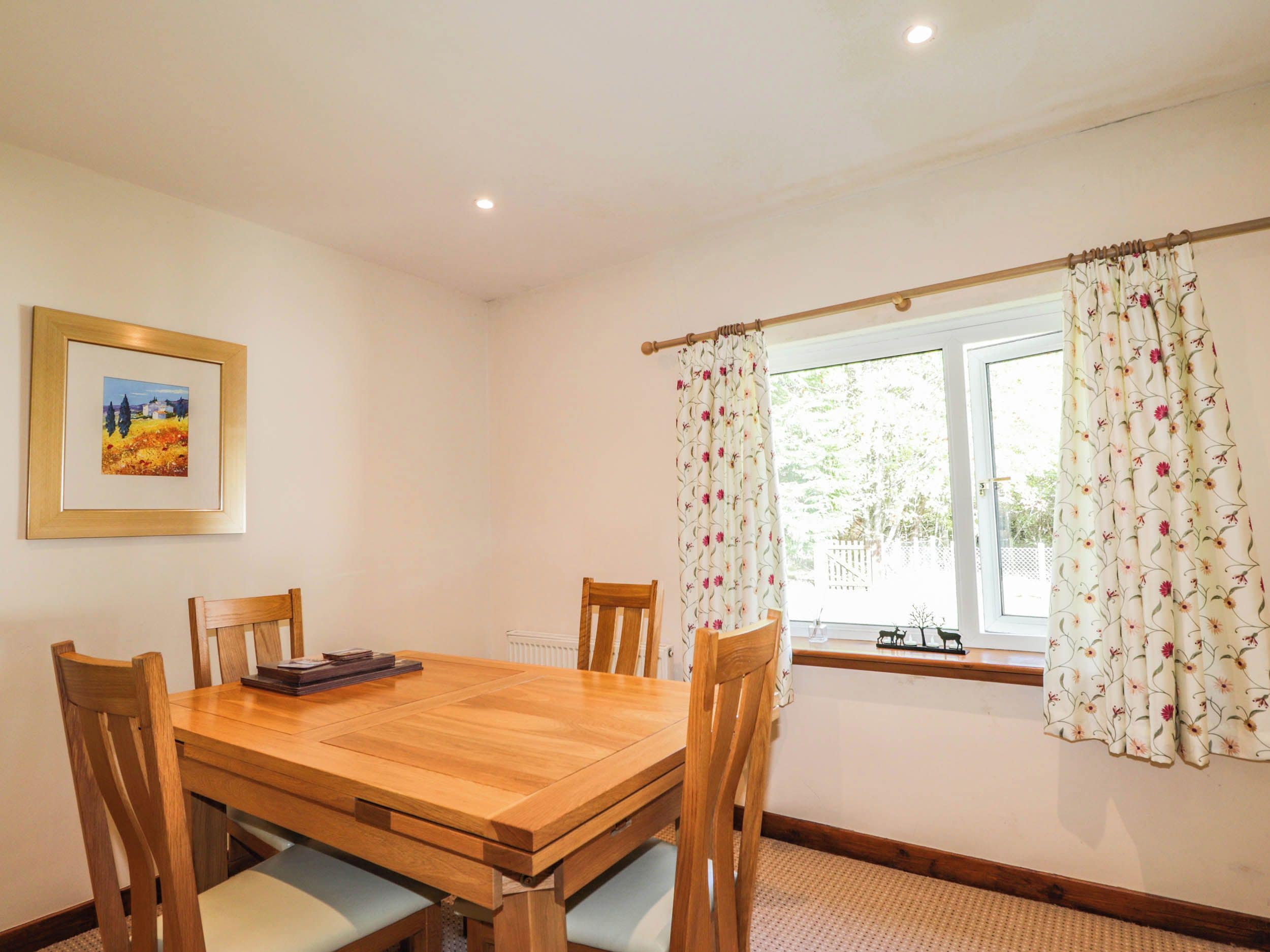 A dining room with a wooden table and chairs, a framed picture on the wall, and a window with floral curtains.
