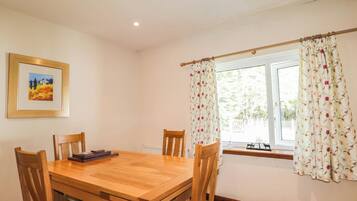 A dining room with a wooden table and chairs, a framed picture on the wall, and a window with floral curtains.