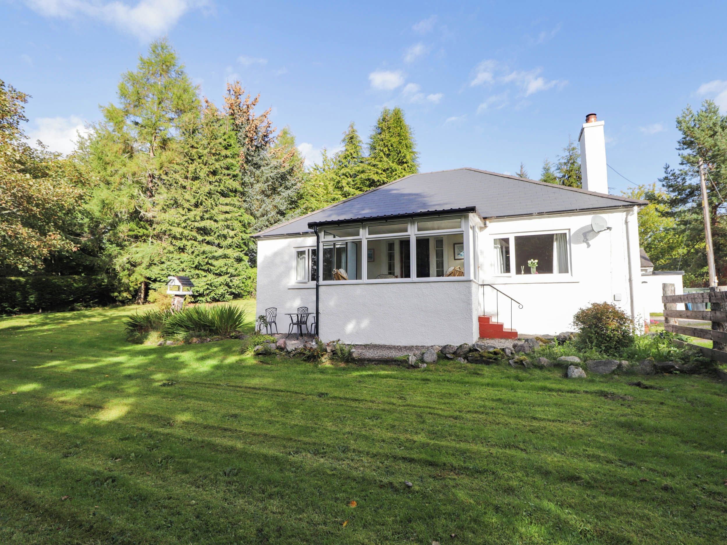 A single-story house with a white exterior, a grey roof, and a chimney. It is surrounded by a well-maintained lawn and dense trees.