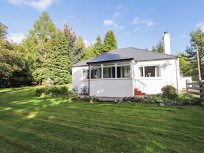 A single-story house with a white exterior, a grey roof, and a chimney. It is surrounded by a well-maintained lawn and dense trees. - St. Aethans (Invershin)