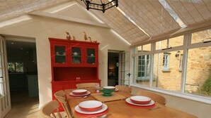A dining area with a wooden table set for four, red cabinets with glass doors, and a view of a stone building through large windows. - Keen Cottage (Chipping Norton)