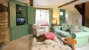 A cozy living room with a green sofa, a floral armchair, and a wooden coffee table. A television is mounted on a green cabinet, and a window with curtains is visible. - Keen Cottage (Chipping Norton)