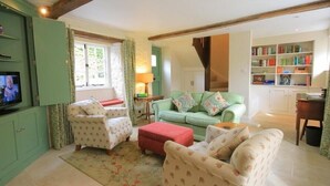 A cozy living room with a green sofa, floral armchairs, a red ottoman, and a television. - Keen Cottage (Chipping Norton)