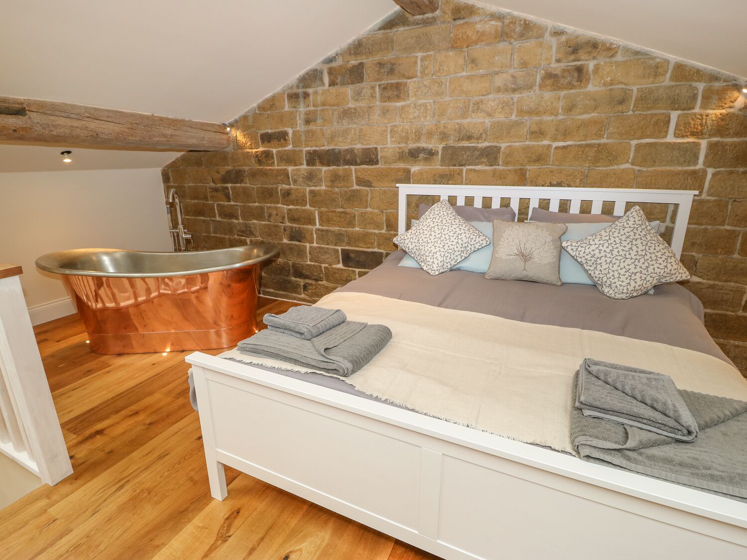 A bedroom with a stone wall, a white bed, grey towels, and a copper bathtub.