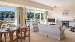 A spacious living room with a dining area, a fireplace, and large windows offering a view of greenery. - 5 Fairhaven (Salcombe)